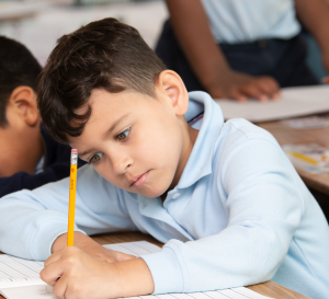 A child with brown hair intently writes with a pencil in a notebook while sitting at a desk in a classroom.