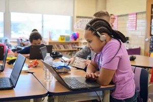 Los estudiantes están sentados en un escritorio del aula usando computadoras portátiles. Un estudiante con una camisa rosa y auriculares se concentra en la pantalla.