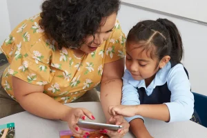 An adult and child are seated at a table, looking at a smartphone together. The adult wears a yellow floral top, and the child is pointing at the screen, engaging with Amplify Desmos Math as part of their interactive mathematics curriculum experience.